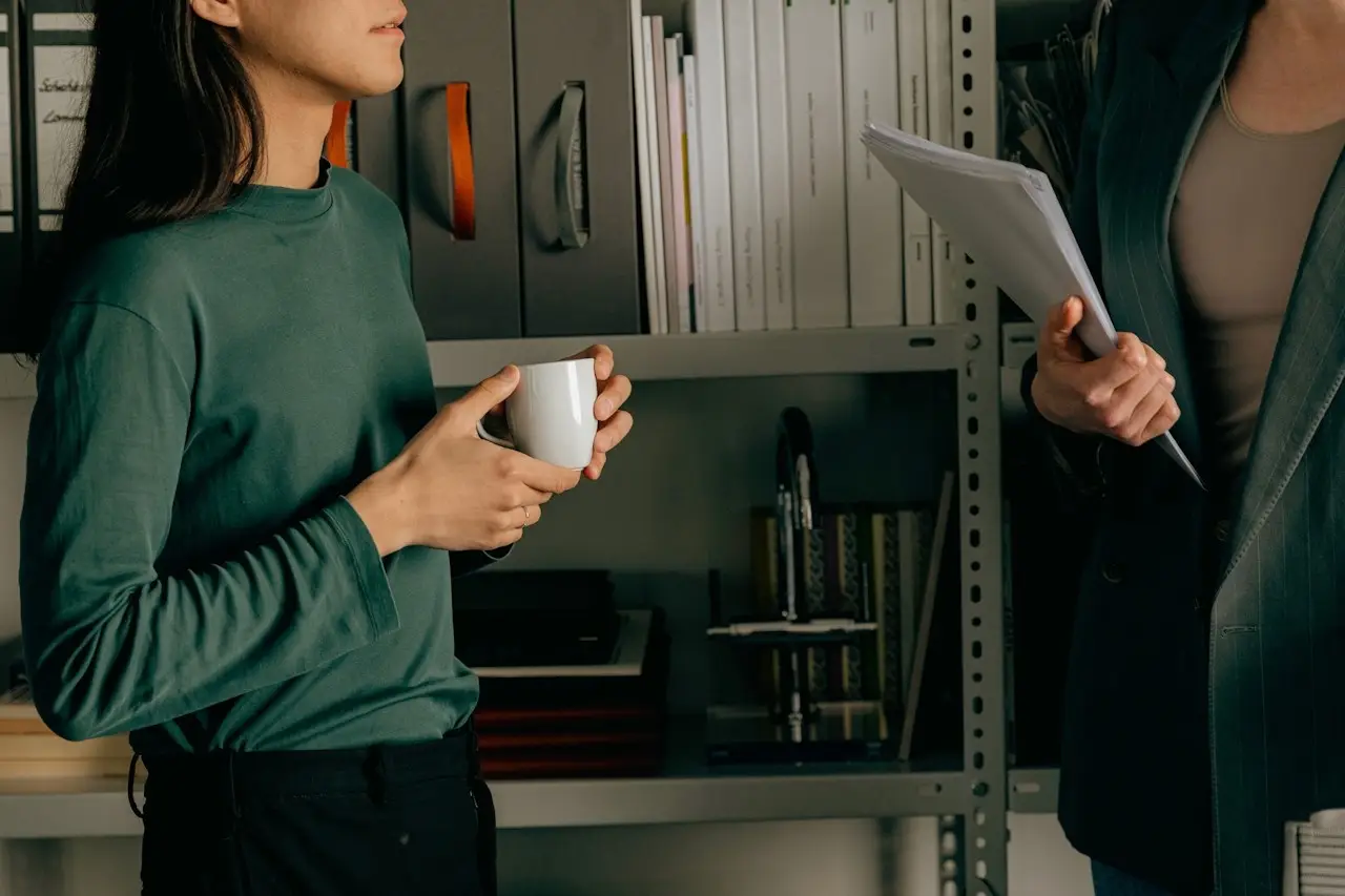 Nahaufnahme einer Hand mit einem weißen Becher in einem Büro; im Hintergrund sind unscharfe Bücherregale zu sehen.