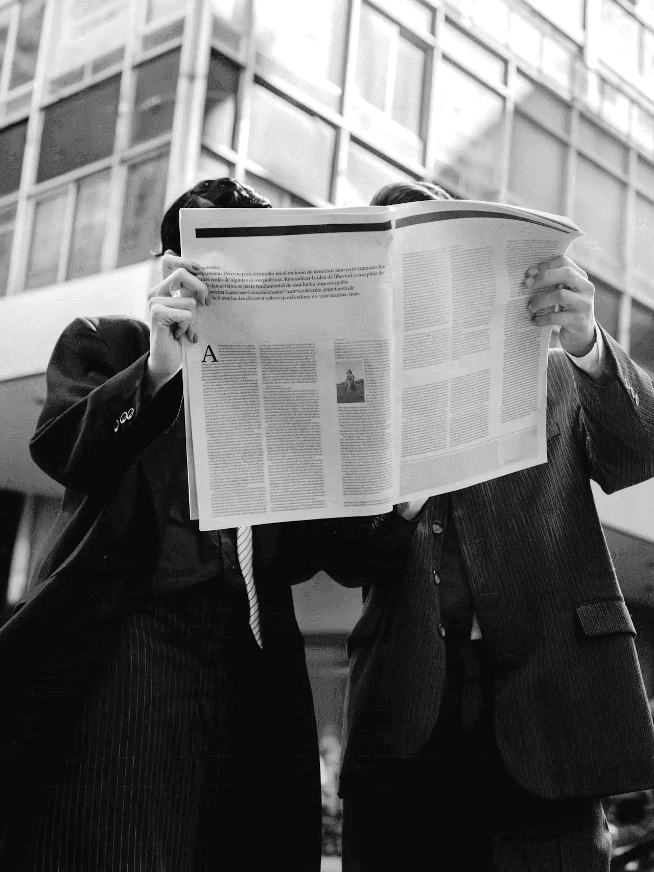 Schwarz-Weiß-Foto von zwei Personen in Geschäftskleidung, die nebeneinander stehen und gemeinsam eine große, aufgeschlagene Zeitung lesen.