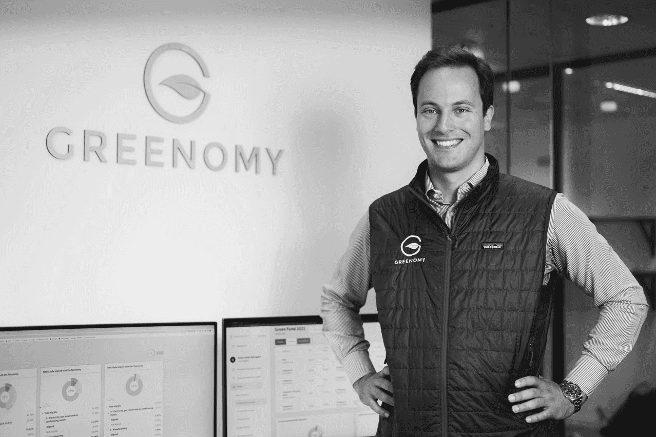 Professional portrait of a man at Greenomy, posing near workstations displaying data dashboards in an office setting.