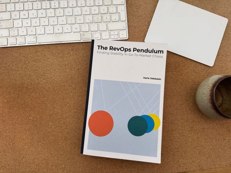 Het boek "The RevOps Pendulum" gecentreerd op een kurken bureau naast een wit computertoetsenbord en trackpad.