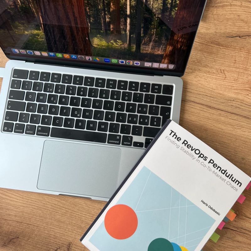 Het boek "The RevOps Pendulum" gecentreerd op een kurken bureau naast een wit computertoetsenbord en trackpad.